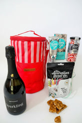 Bottle of sparkling wine, red striped cooler bag, and assorted snacks on a textured surface. Gift hamper for her