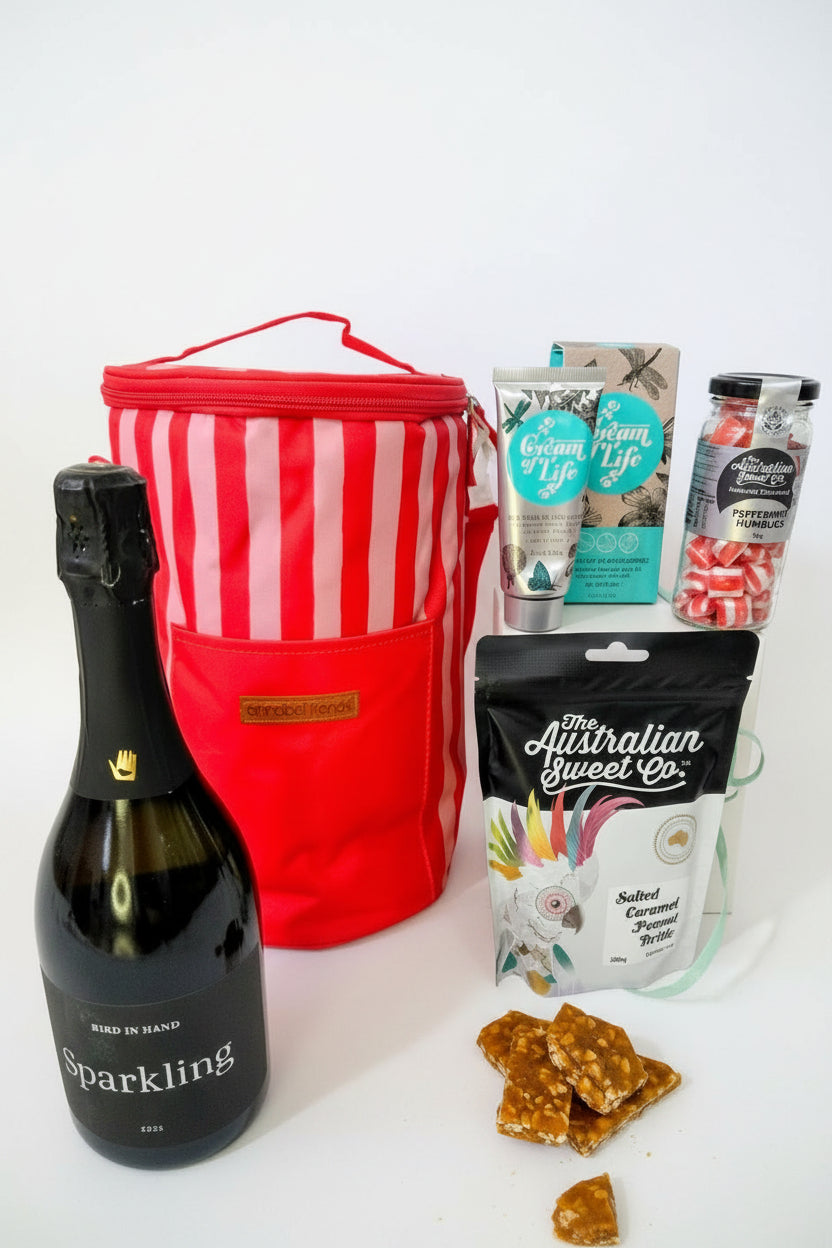 Bottle of sparkling wine, red striped cooler bag, and assorted snacks on a textured surface. Gift hamper for her