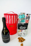 Bottle of sparkling wine, red striped cooler bag, and assorted snacks on a textured surface. Gift hamper for her