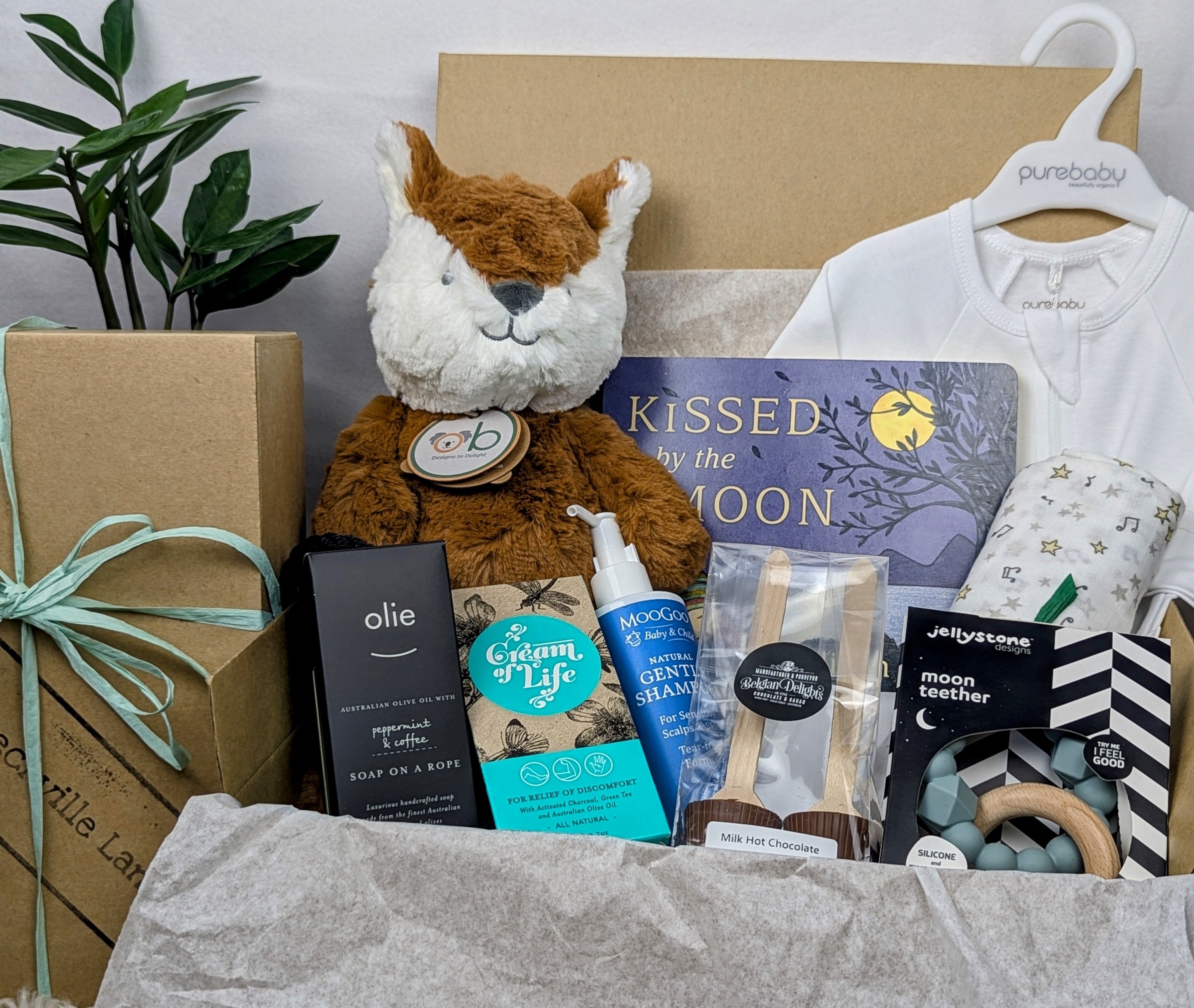 A gift box containing a variety of baby-related items including a soft OB Designs Frankie Fox toy, growsuit, moogoo shampoo, teether, book, muslin, hot chocolate spoons, soap on a rope, and luxury cream, all beautifully arranged in a kraft box with raffia ribbon. Baby boy gift, baby boy hamper, corporate maternity hamper. 