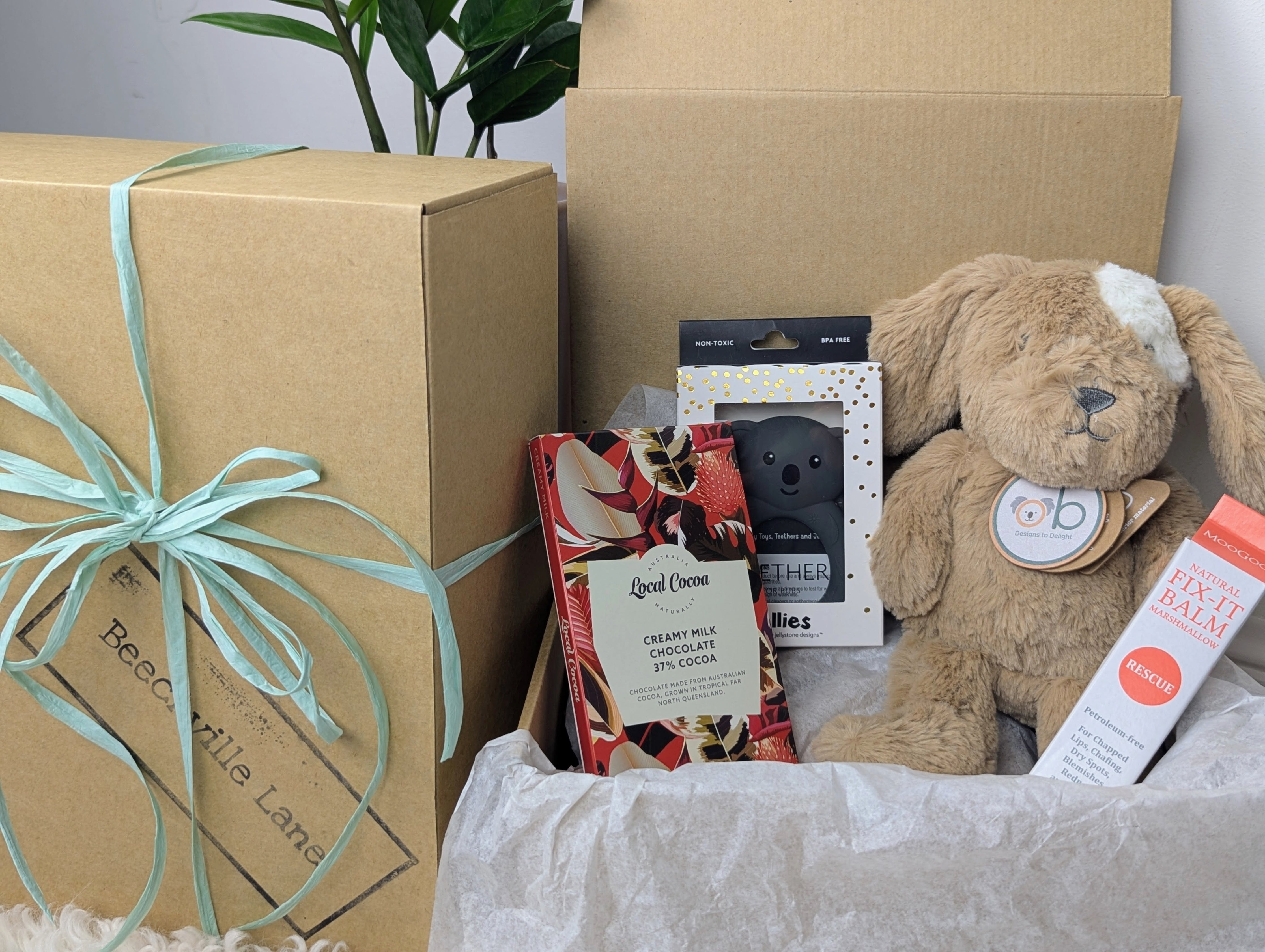 A gift box open to reveal its contents, which include a caramel-colored dog soft toy, a small jar of balm, a teether, and a chocolate bar. OB Designs, MooGoo, Baby Hamper Australia. 