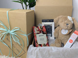A gift box open to reveal its contents, which include a caramel-colored dog soft toy, a small jar of balm, a teether, and a chocolate bar. OB Designs, MooGoo, Baby Hamper Australia. 
